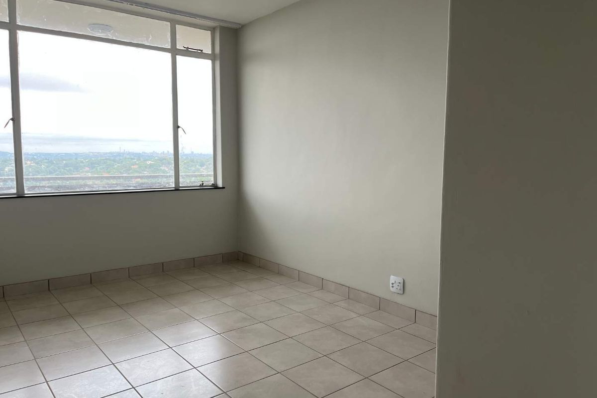 An empty bedroom featuring neutral tiled flooring, large windows with natural light, and a view of the surrounding area.