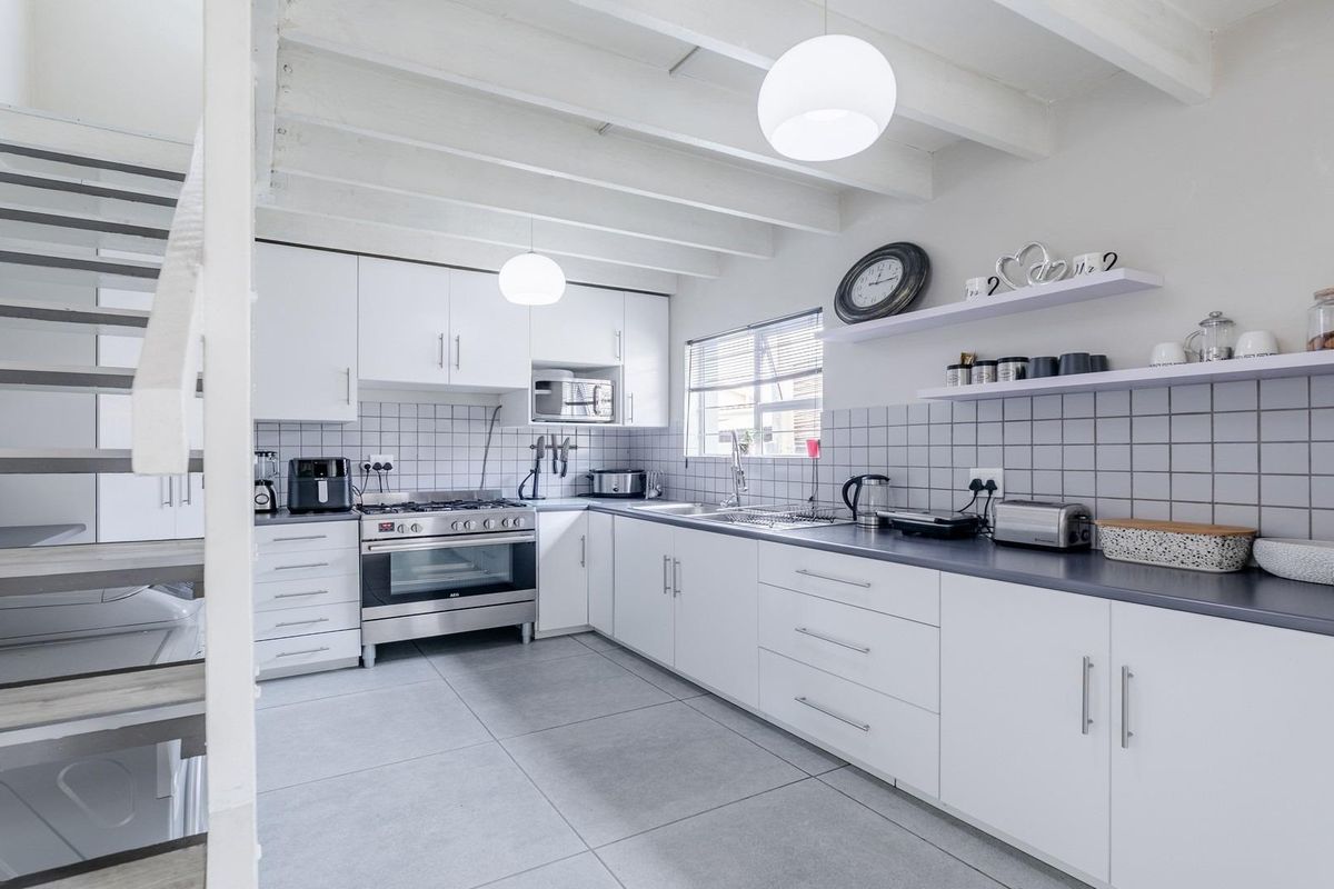 Modern kitchen featuring white cabinetry, tiled backsplash, dark countertops, stainless steel oven, and exposed white ceiling beams with an adjacent staircase.