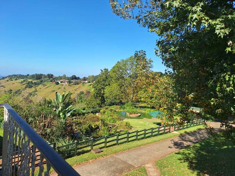 Scenic elevated view overlooking a lush landscaped garden, pond, and surrounding hillside landscape from a private balcony with a wrought iron balustrade.