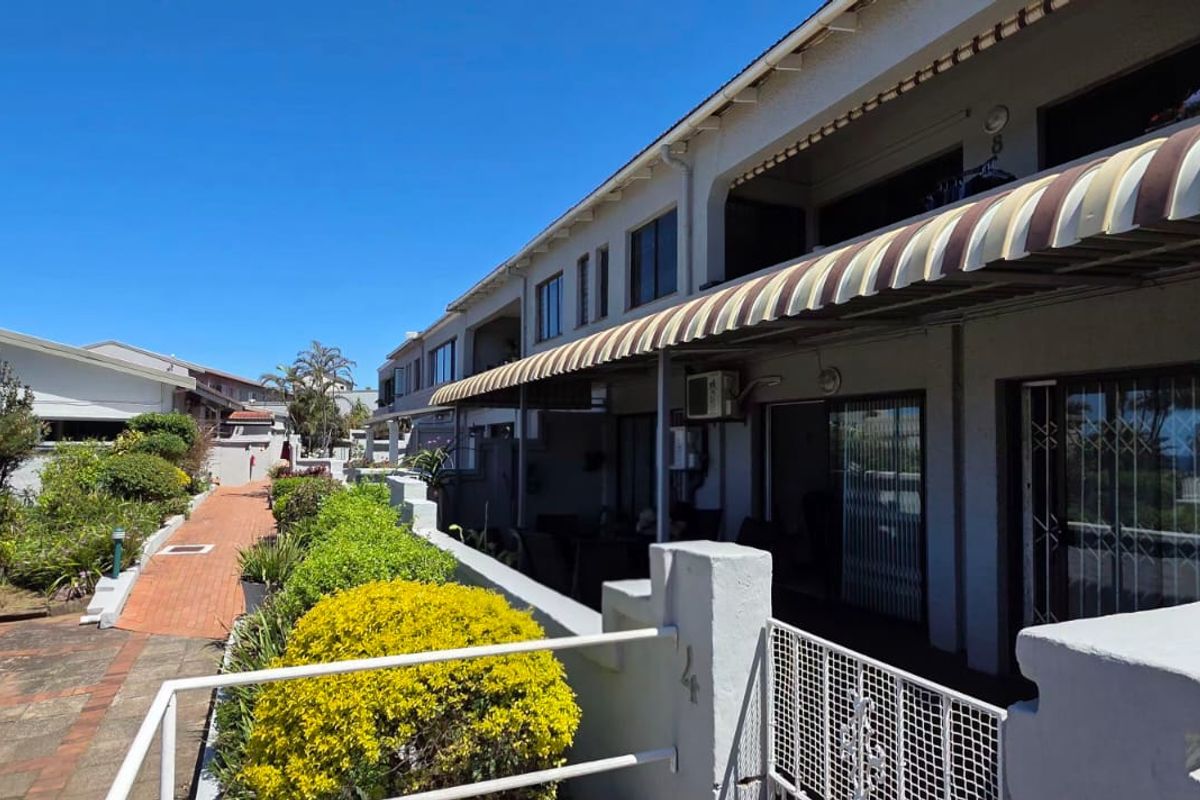 Rear exterior view of the apartment building featuring a paved walkway, landscaped garden area, and covered patio space with striped awnings.