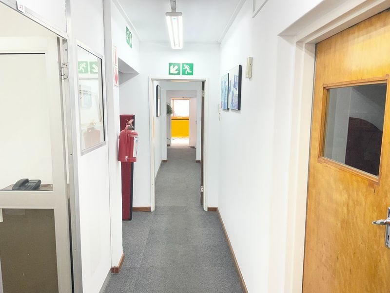 A functional industrial office hallway featuring carpeted flooring, emergency exit signage, and a timber door with a vision panel.