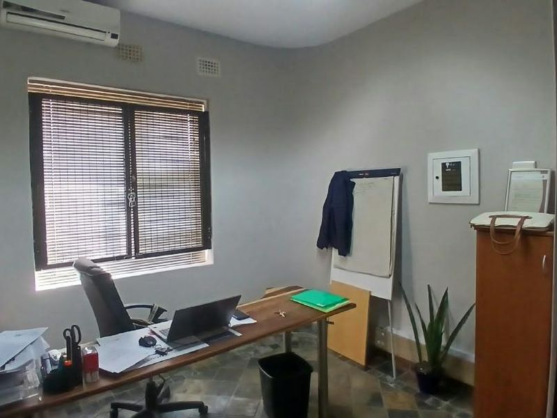 A functional office space featuring tiled flooring, a wall-mounted air conditioning unit, and window blinds, providing a practical work environment.
