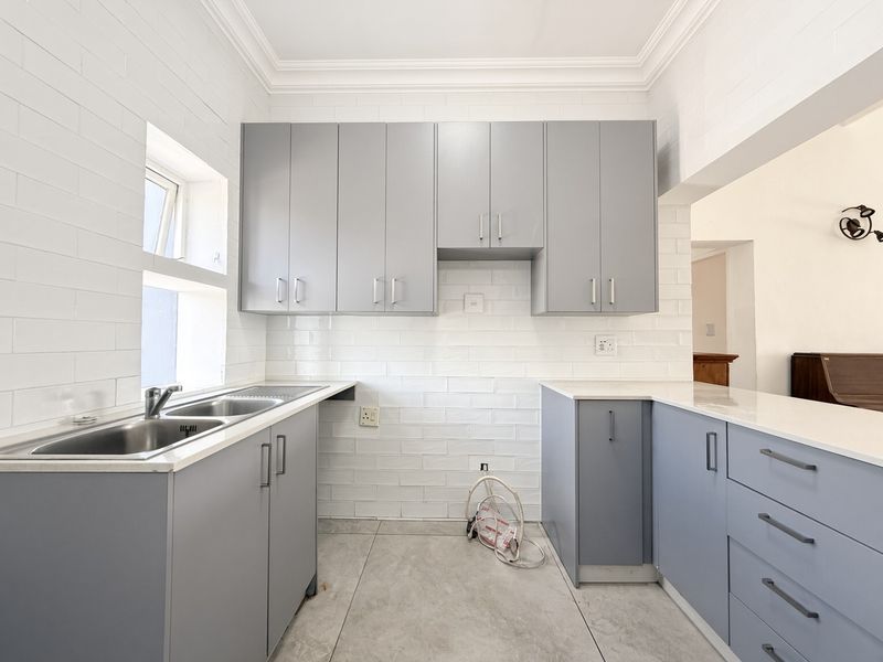 A clean, modern kitchen featuring grey built-in cabinetry, white subway tile backsplash, stone countertops, and a stainless steel double sink.