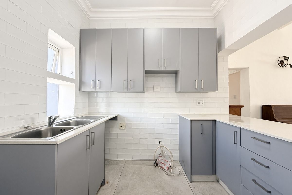 A clean, modern kitchen featuring grey built-in cabinetry, white subway tile backsplash, stone countertops, and a stainless steel double sink.