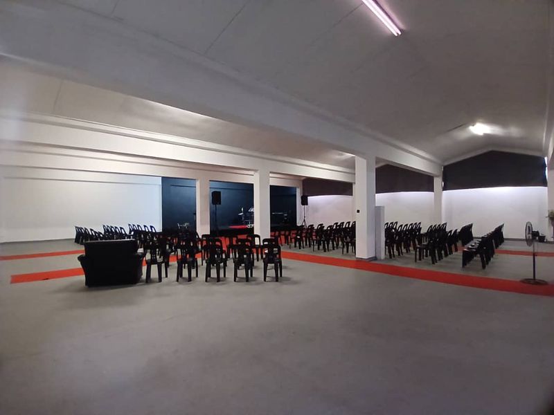Spacious open-plan event hall or clubhouse featuring a raised stage area, neutral painted walls, and industrial-style fluorescent lighting.