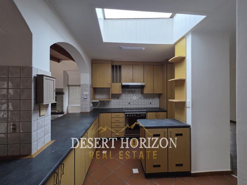 A functional kitchen featuring wooden cabinetry, dark countertops, a central skylight providing natural light, and terracotta floor tiles.