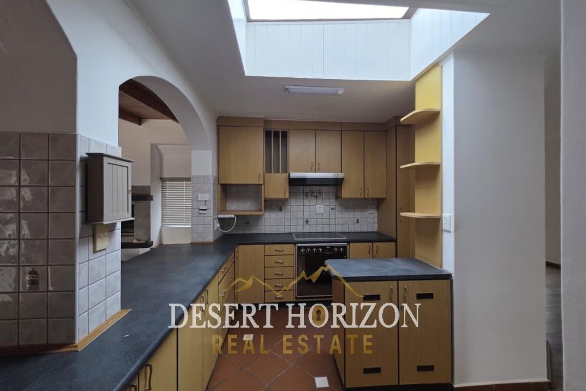 A functional kitchen featuring wooden cabinetry, dark countertops, a central skylight providing natural light, and terracotta floor tiles.