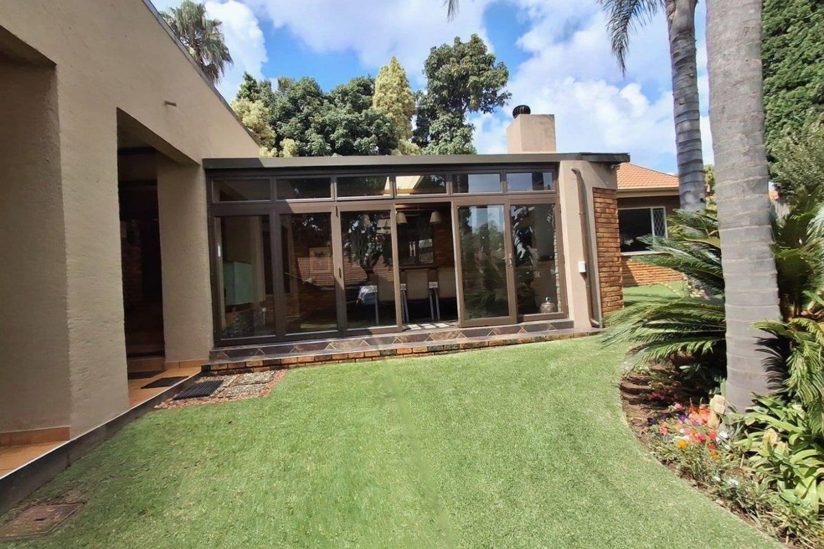 A well-maintained rear garden featuring a modern enclosed patio with large aluminium sliding doors and a brick chimney, set against a lush lawn.