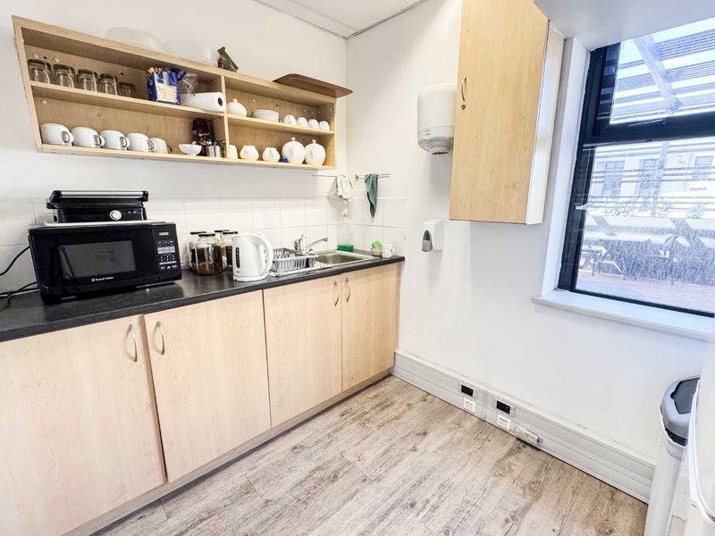 A functional kitchenette featuring light wood-finish cabinetry, dark countertops, a stainless steel sink, and open shelving, suitable for office or staff use.