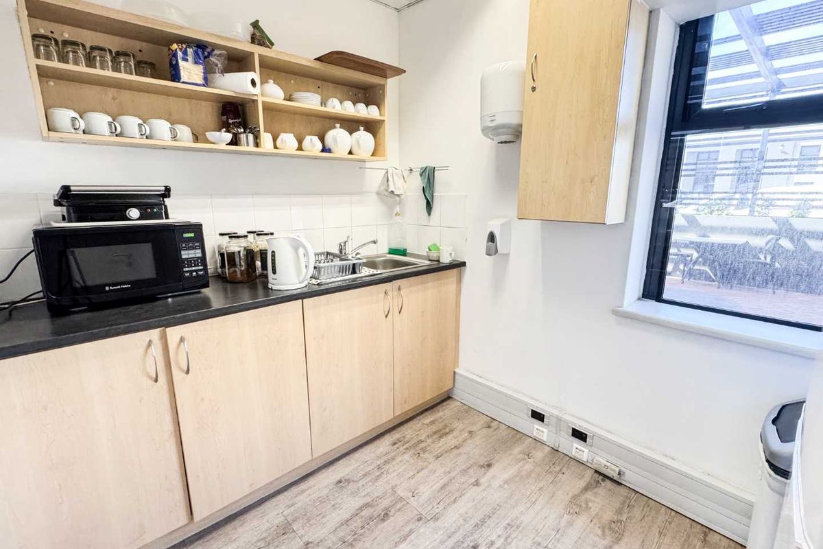 A functional kitchenette featuring light wood-finish cabinetry, dark countertops, a stainless steel sink, and open shelving, suitable for office or staff use.