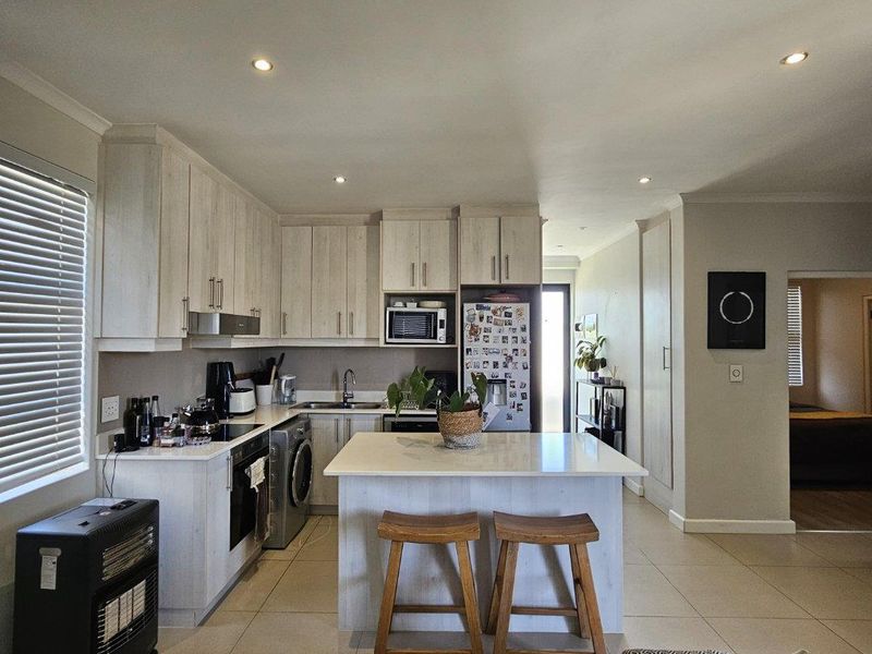 Modern open-plan kitchen featuring light wood-finish cabinetry, stone countertops, a central island, and integrated appliances, seamlessly flowing into the living area.