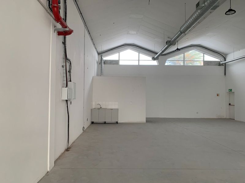 Spacious open-plan retail or industrial unit featuring high vaulted ceilings, exposed ductwork, concrete flooring, and a functional kitchenette area.