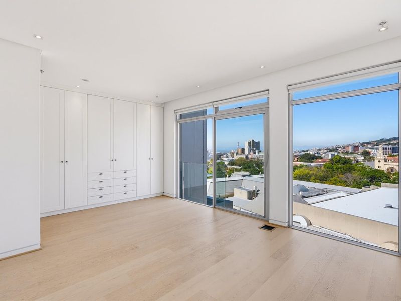 A bright, modern bedroom featuring light hardwood flooring, extensive built-in cabinetry, and large sliding glass doors offering expansive city and landscape views.