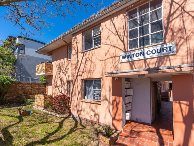 Exterior view of Winton Court apartment building featuring a stucco facade, tiled entrance, and communal mailboxes with surrounding garden area.
