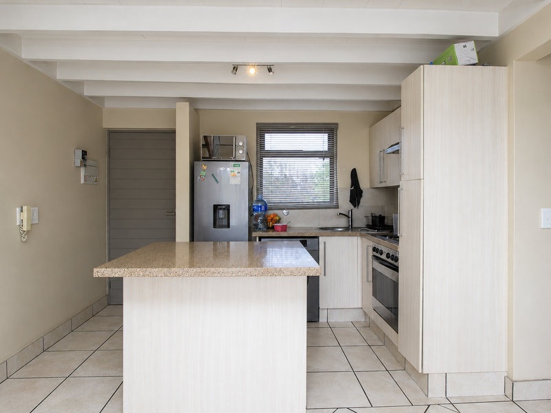 A functional kitchen featuring a central island, light-toned cabinetry, built-in oven with hob, and tiled flooring under an exposed beam ceiling.