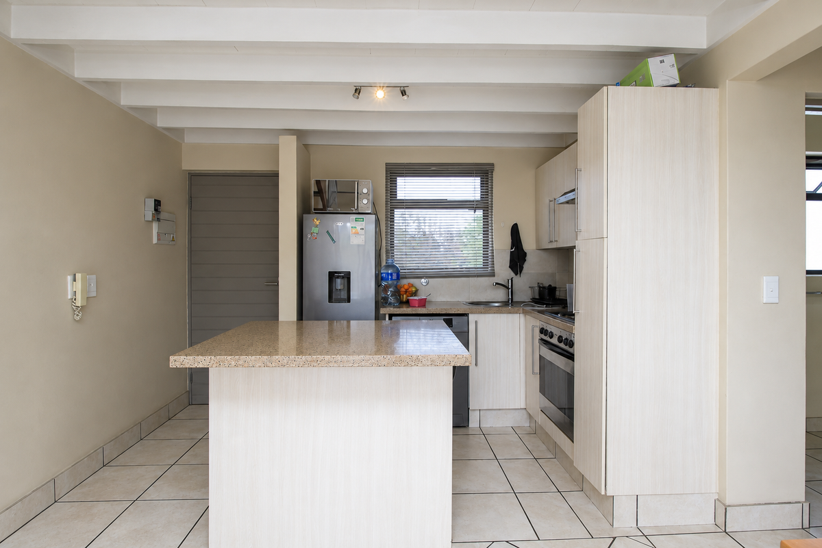 A functional kitchen featuring a central island, light-toned cabinetry, built-in oven with hob, and tiled flooring under an exposed beam ceiling.