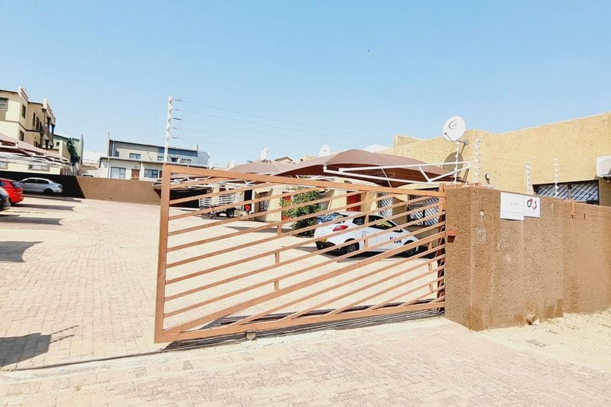 Front exterior view of the townhouse complex featuring a paved driveway, secure sliding gate, carport parking, and perimeter electric fencing.