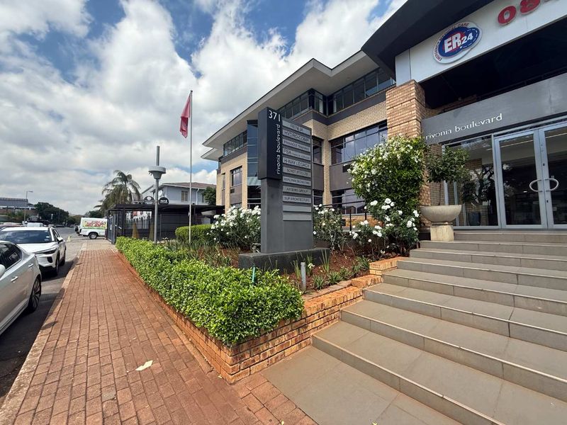 Front exterior view of a commercial office building on Rivonia Boulevard, featuring a paved walkway, landscaped garden, and prominent business signage pylon.