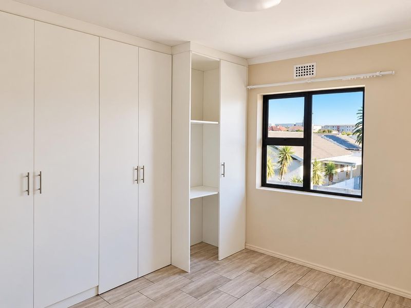 A bright, empty bedroom featuring modern white built-in cupboards, wood-look tiled flooring, and a window with an exterior view.