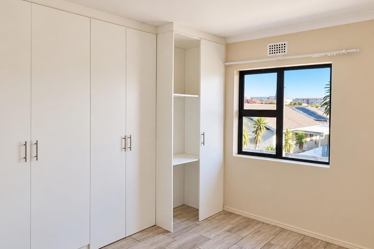 A bright, empty bedroom featuring modern white built-in cupboards, wood-look tiled flooring, and a window with an exterior view.