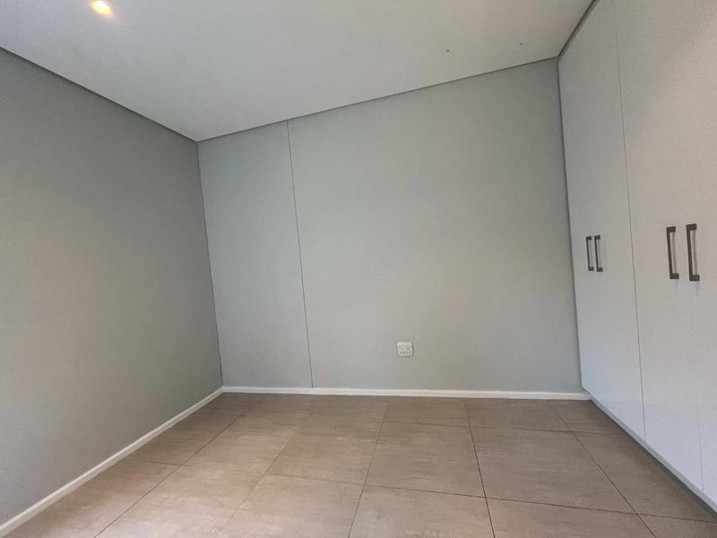 A clean, empty bedroom featuring neutral grey walls, large format floor tiles, and modern white built-in cupboards with integrated storage.