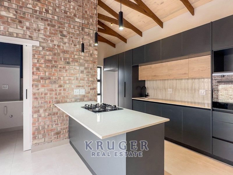 Modern kitchen featuring exposed brick walls, timber ceiling beams, a central island with gas hob, and sleek dark cabinetry with integrated appliances.