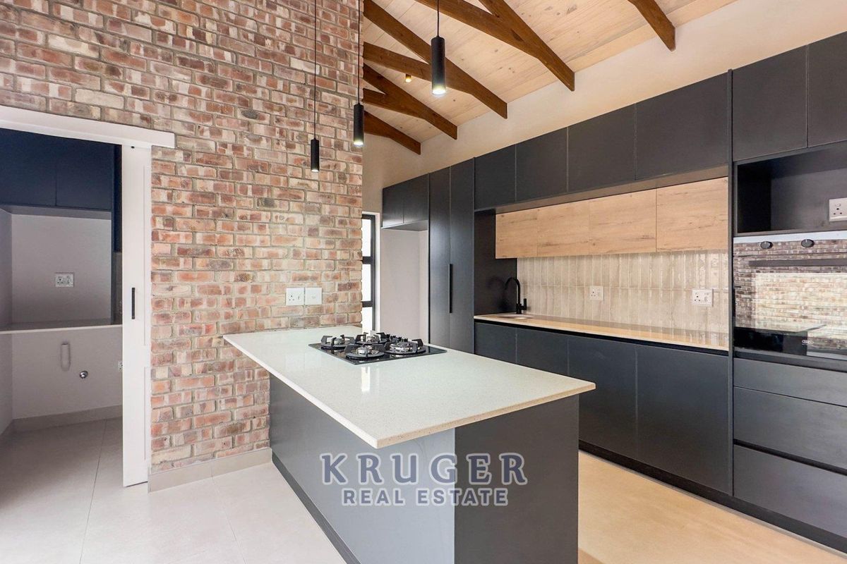 Modern kitchen featuring exposed brick walls, timber ceiling beams, a central island with gas hob, and sleek dark cabinetry with integrated appliances.