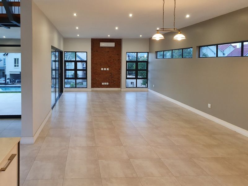 Spacious open-plan living area featuring tiled floors, an exposed brick feature wall, split-system air conditioning, and large windows providing ample natural light.
