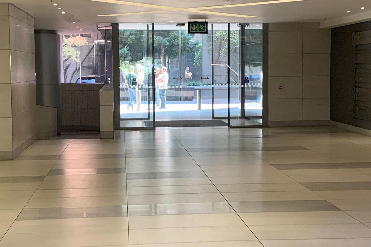A spacious and modern office building entrance lobby featuring polished tiled flooring, recessed ceiling lighting, and large glass sliding doors.