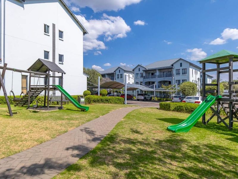 A well-maintained communal garden area featuring a paved walkway, landscaped lawns, and playground equipment set against the apartment complex buildings.