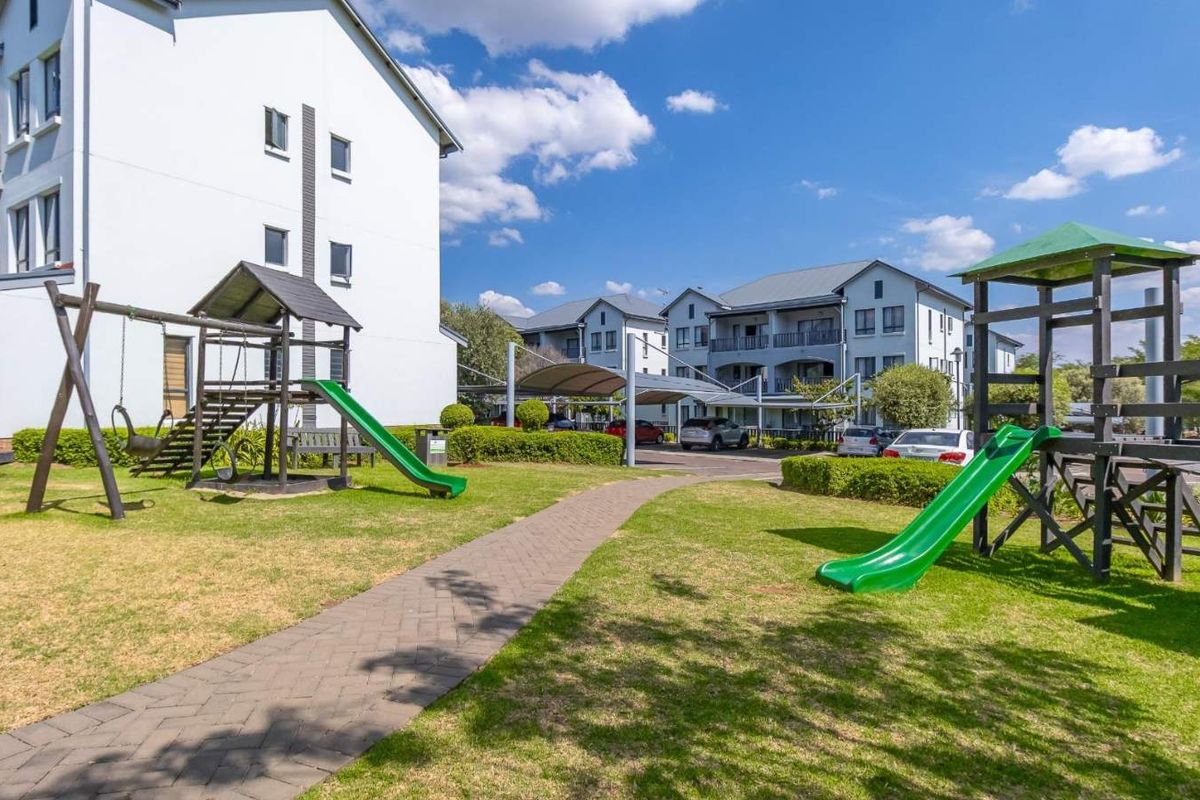 A well-maintained communal garden area featuring a paved walkway, landscaped lawns, and playground equipment set against the apartment complex buildings.