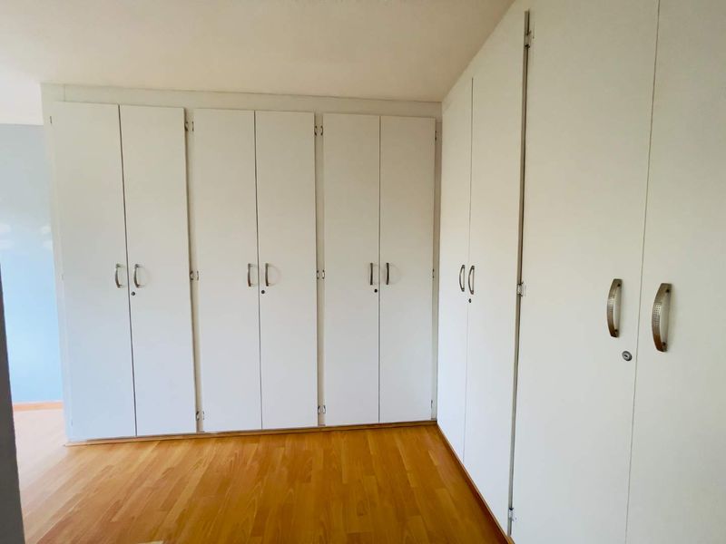 A bright bedroom featuring extensive white built-in cupboards and warm-toned laminate flooring, offering ample storage space.