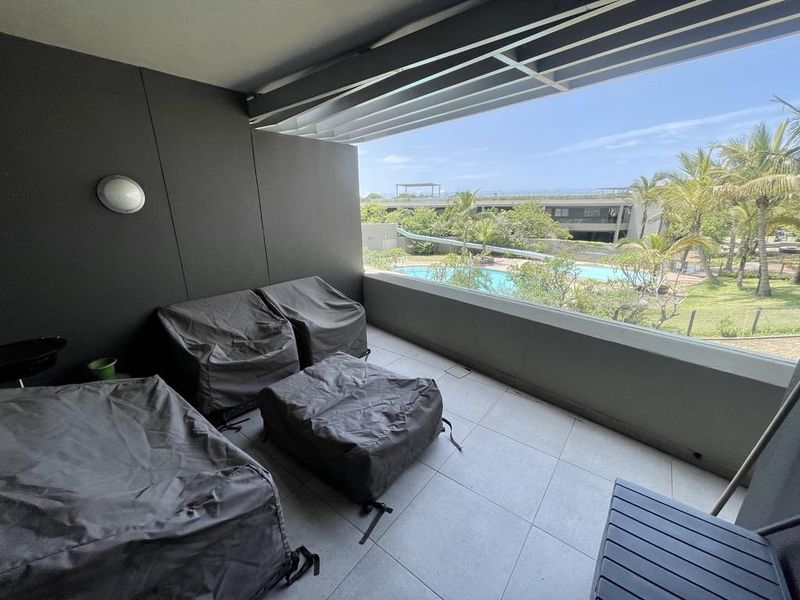 A modern covered balcony featuring tiled flooring, a glass balustrade, and a louvred roof, offering pleasant views of the complex pool and garden area.