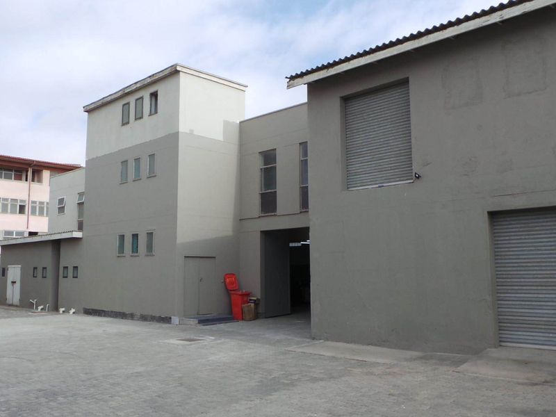 Exterior view of a commercial retail property featuring concrete paving, roller shutter doors, and multi-level building structure.