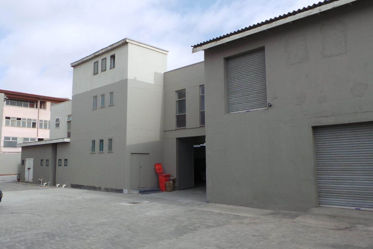 Exterior view of a commercial retail property featuring concrete paving, roller shutter doors, and multi-level building structure.