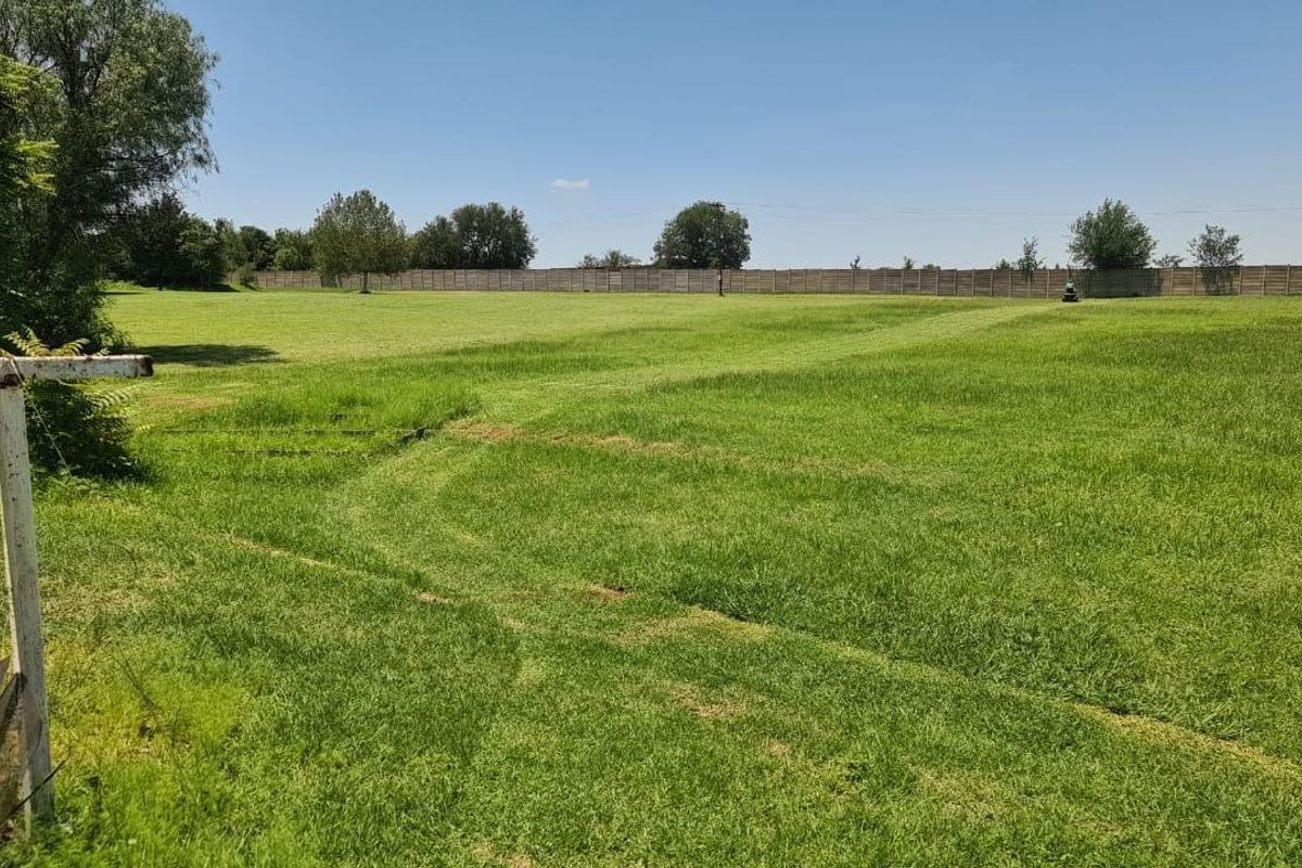 A wide, open agricultural land plot featuring expansive green grass, mature trees, and a perimeter fence boundary.