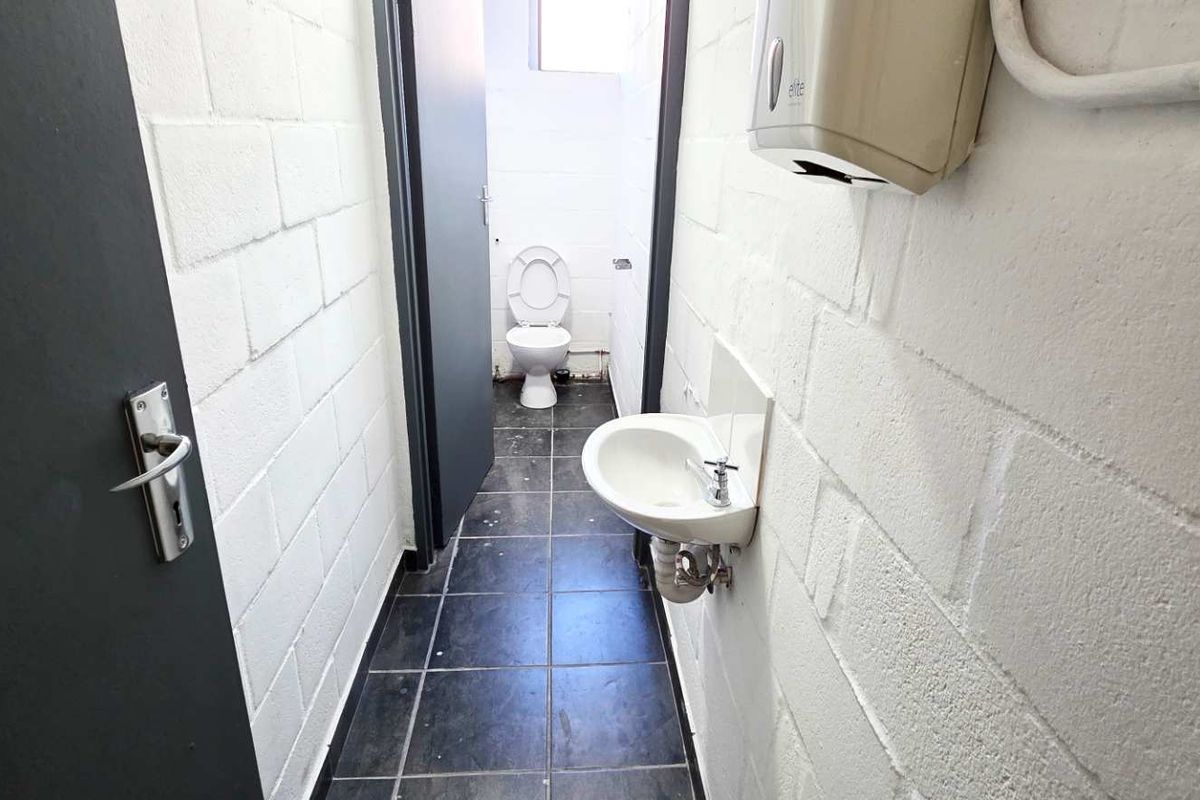 A functional industrial restroom featuring a wall-mounted basin, toilet, and tiled flooring with exposed brickwork walls.