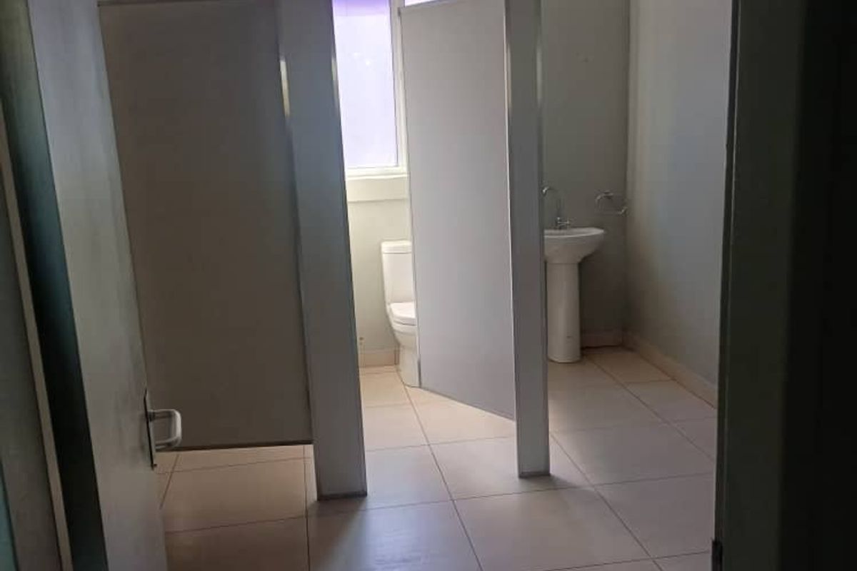 A functional office bathroom featuring partitioned toilet cubicles, a pedestal basin, tiled flooring, and a suspended ceiling.