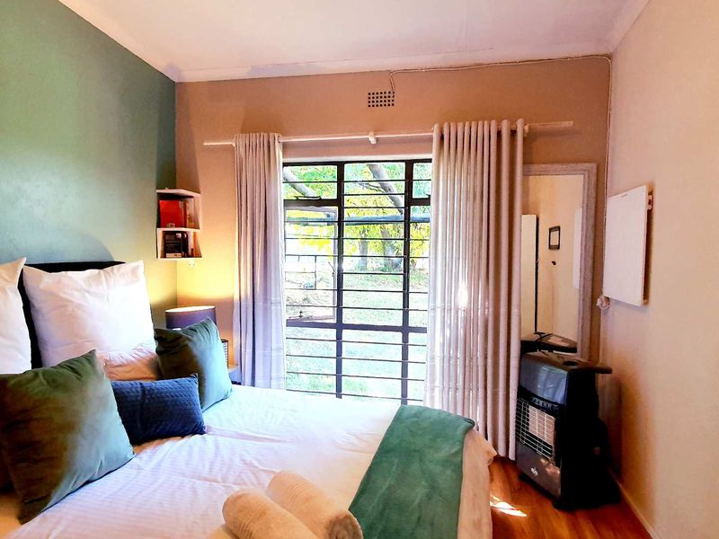 A cozy bedroom featuring a large window with security bars, wooden flooring, and a feature wall, providing natural light and garden views.