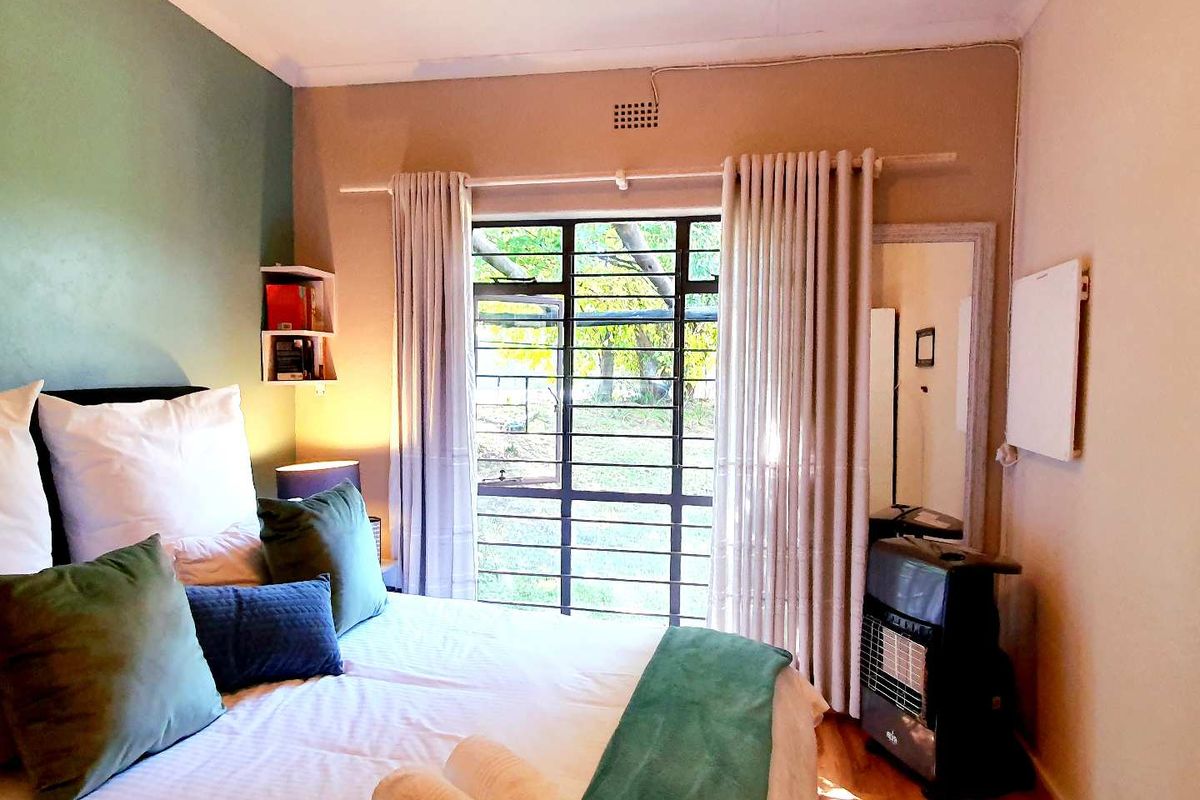 A cozy bedroom featuring a large window with security bars, wooden flooring, and a feature wall, providing natural light and garden views.