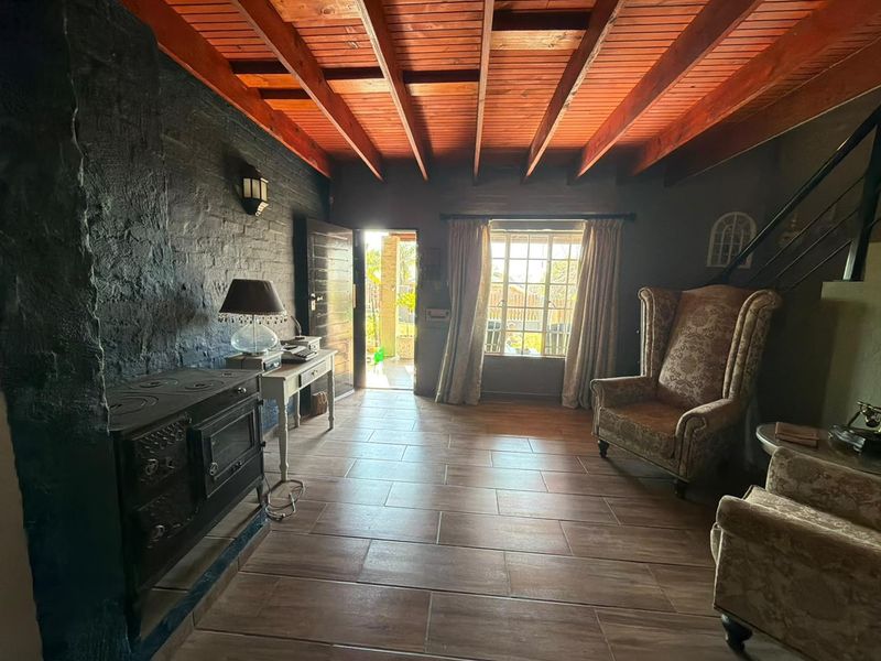 A rustic living room featuring exposed timber beam ceilings, dark painted brick walls, tiled flooring, and a traditional cast iron wood burning stove.