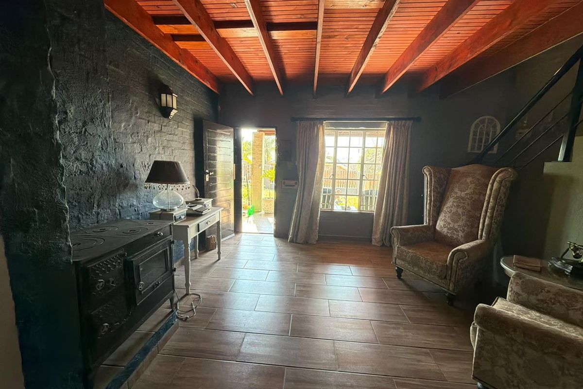 A rustic living room featuring exposed timber beam ceilings, dark painted brick walls, tiled flooring, and a traditional cast iron wood burning stove.
