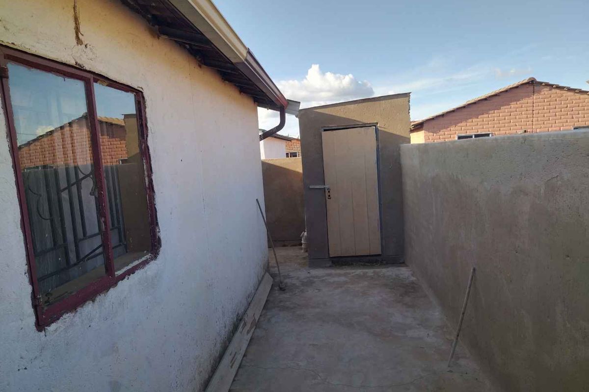 A view of the rear exterior area featuring a concrete pathway, a small utility structure, and perimeter boundary walls.