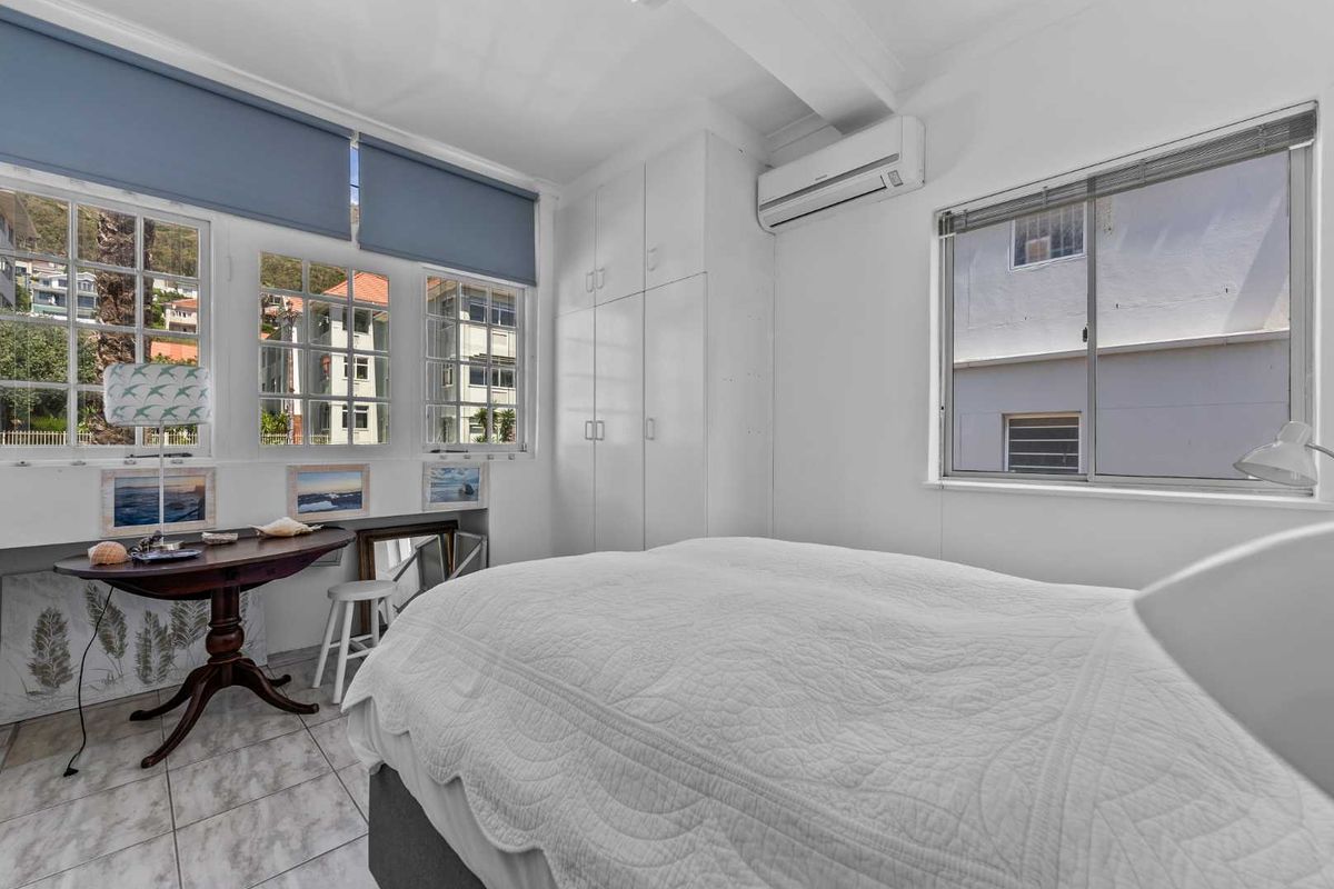 A bright bedroom featuring built-in cupboards, tiled flooring, and a wall-mounted air conditioning unit, with large windows providing natural light.