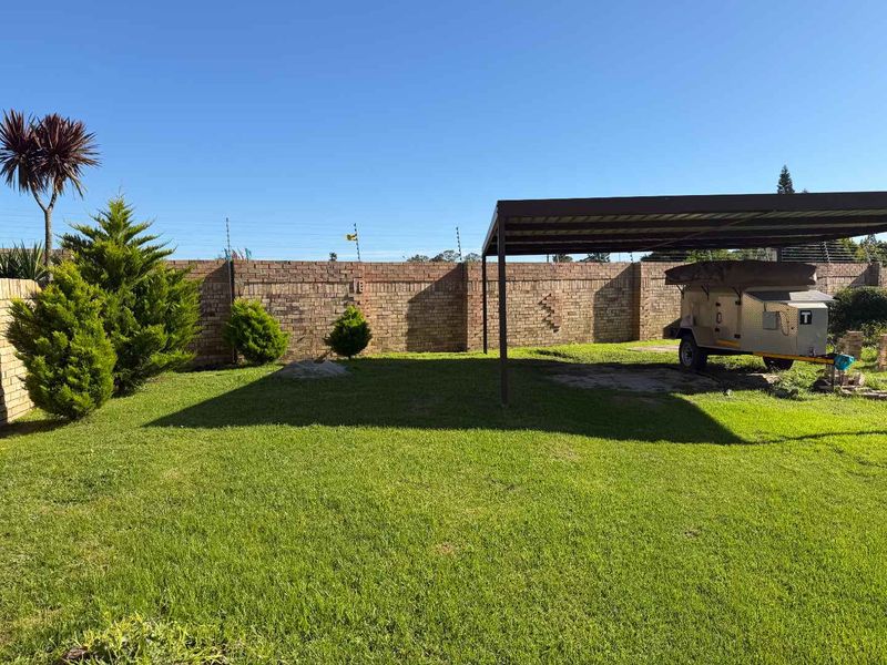 A spacious rear garden featuring a lawn, a metal carport structure, and a brick boundary wall equipped with electric fencing.