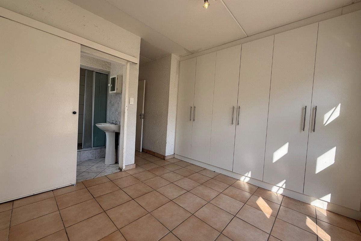 A vacant bedroom featuring built-in cupboards, tiled flooring, and an attached en-suite bathroom with a shower and pedestal basin.