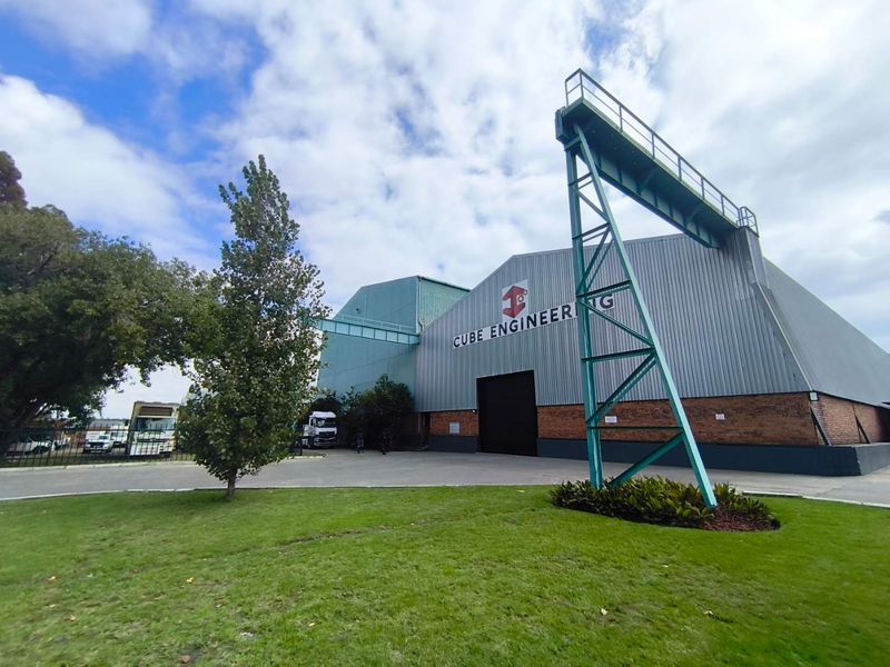 Front view of an industrial facility featuring metal cladding, brick base, and an external gantry crane structure set within a landscaped yard.