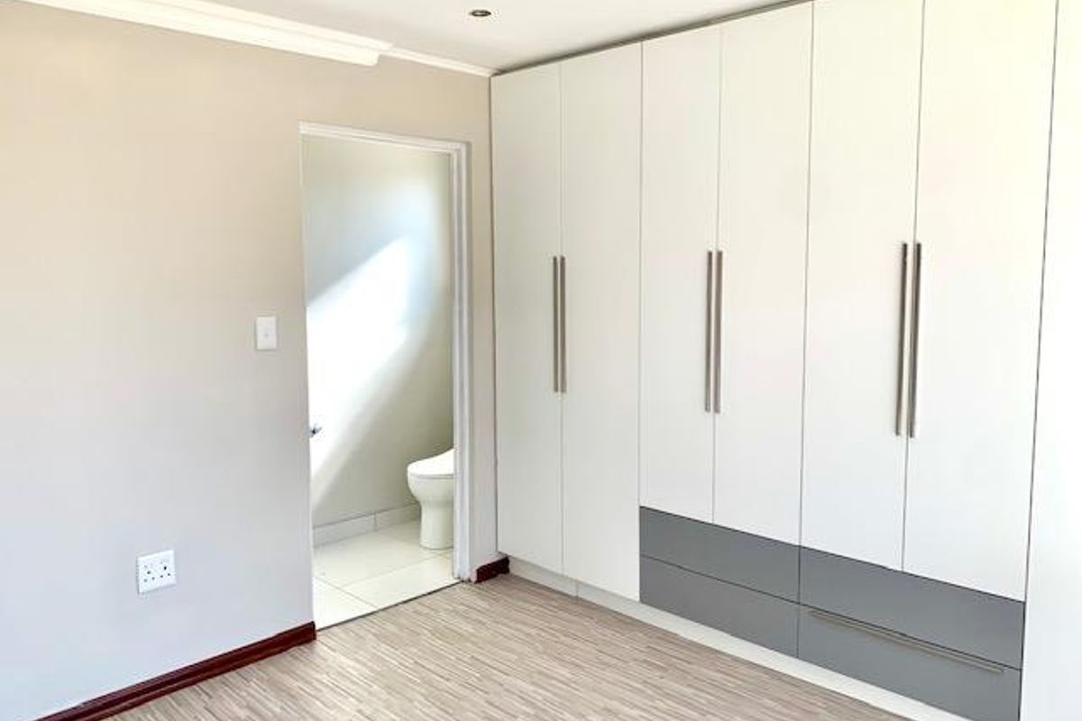 A bright, empty bedroom featuring modern built-in cupboards with a two-tone finish, laminate flooring, and an adjoining en-suite bathroom.