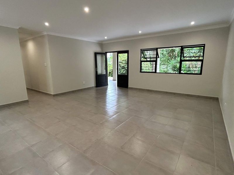 A spacious, empty living area featuring neutral tiled flooring, recessed downlighting, and black-framed windows and doors leading to the exterior.