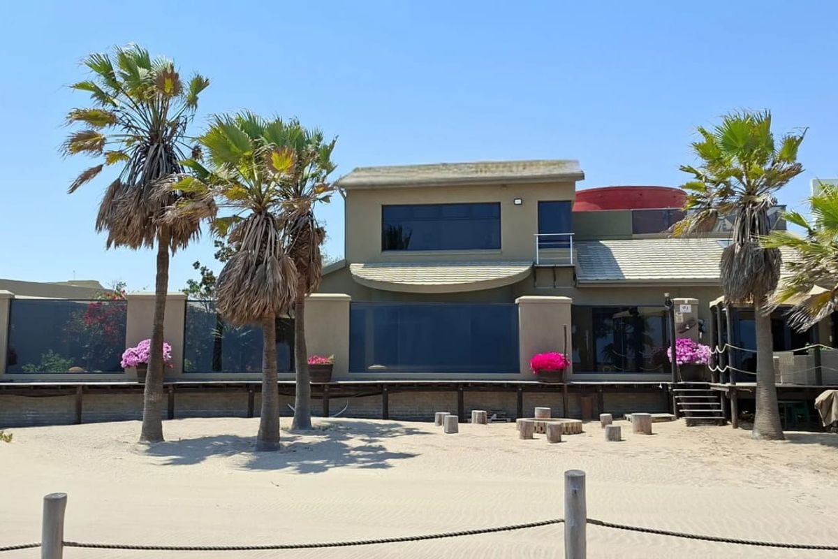 Rear view of a beach house featuring a raised wooden deck, large glass windows, and direct access to the sandy beach area.